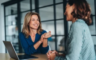 Two women talk across a desk with a laptop during a trustee planning meeting in a modern office