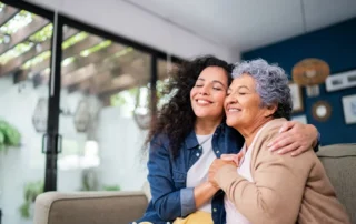 Daughter hugs her mother on sofa, showing what to consider before moving your aging parents at home.