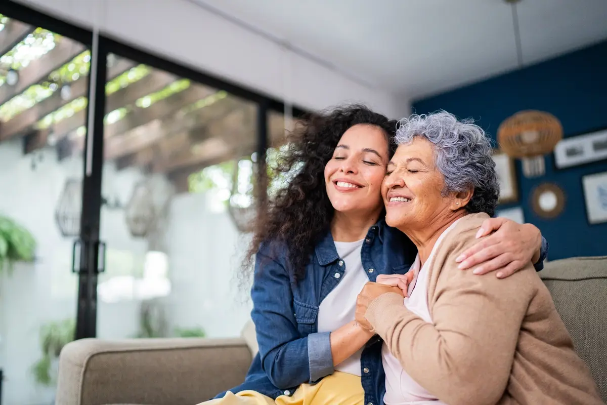 Daughter hugs her mother on sofa, showing what to consider before moving your aging parents at home.