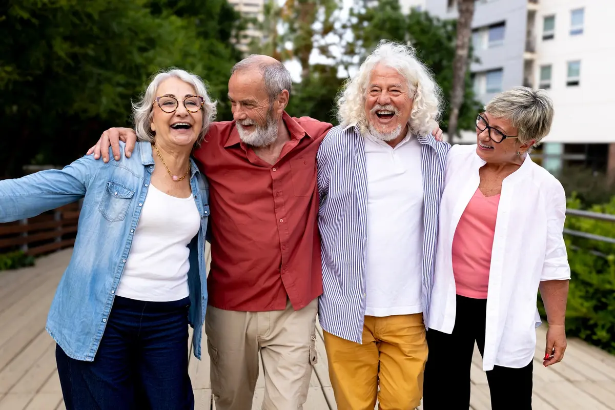 Four older friends walk arm in arm outdoors, smiling as they plan retirement at 65 or 85