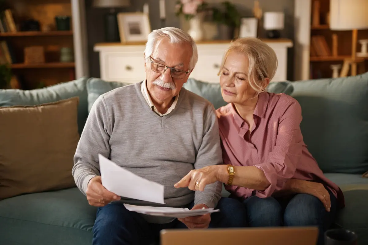 Senior couple reviews trustee papers on a couch at home, looking over documents beside a laptop