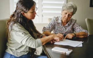Woman helps senior sign power of attorney documents at a table while reviewing paperwork together