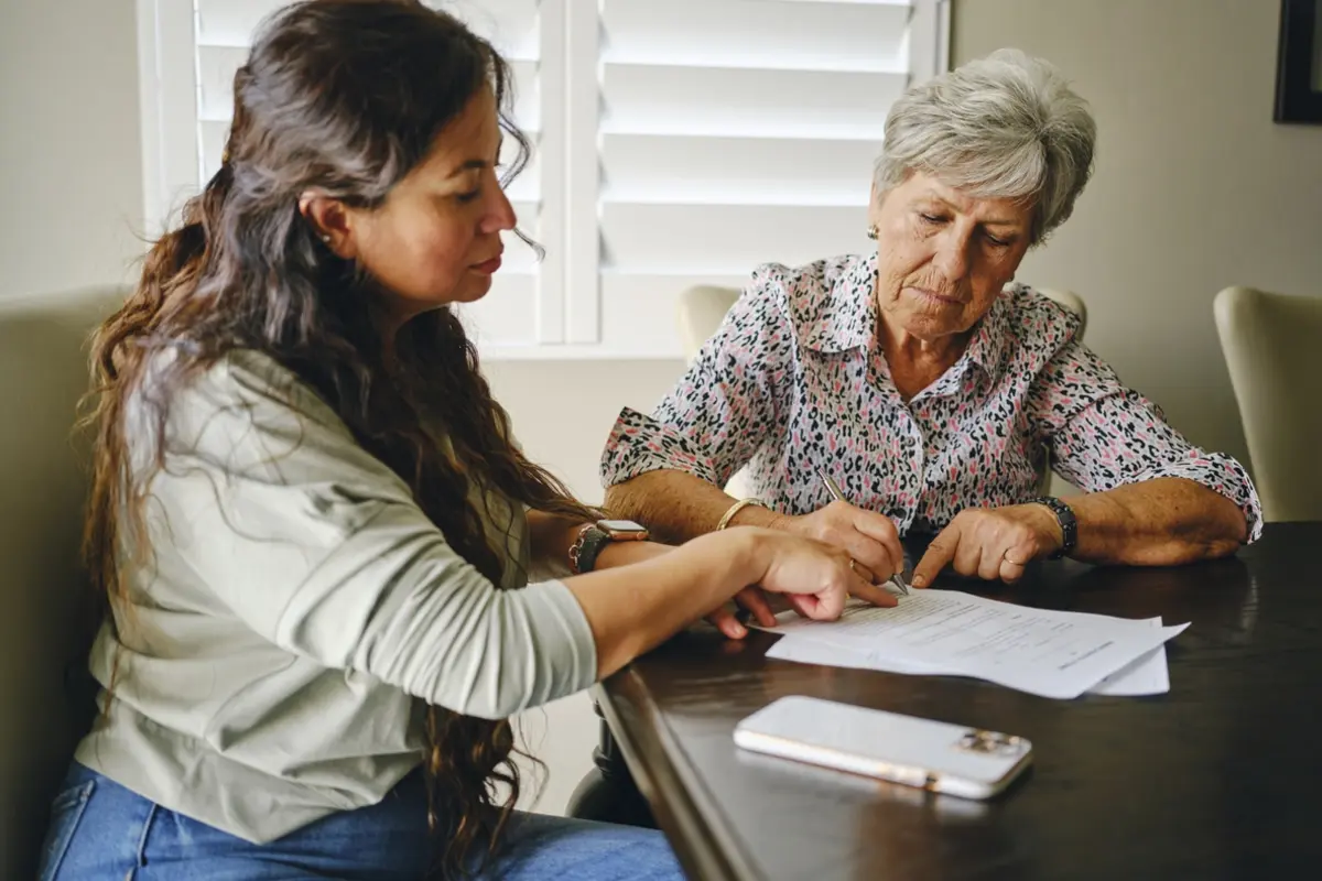 Woman helps senior sign power of attorney documents at a table while reviewing paperwork together